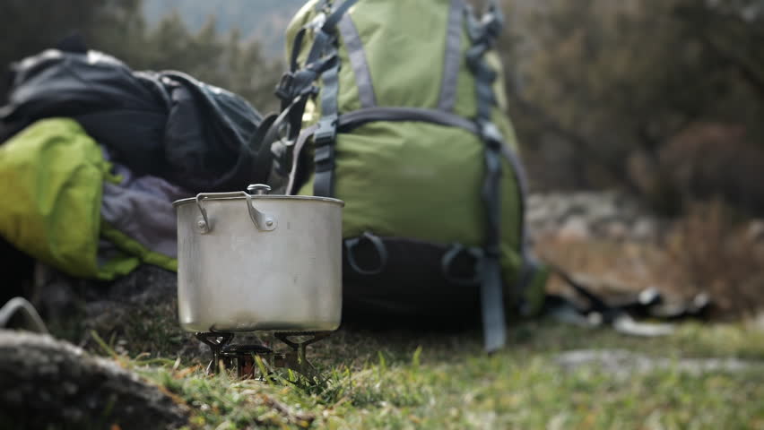 A metal pot for mountain hiking stands on a gas burner on a rock with mountains in the background. A metal pot of boiling water sits on the burner against a backdrop of mountains.