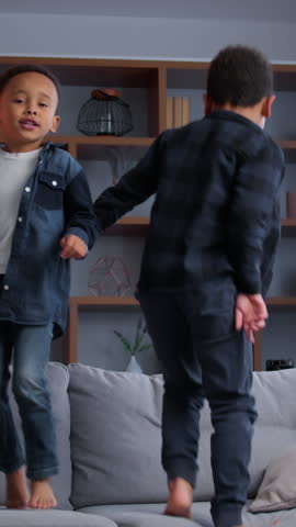 Two little African American kids jumping on couch at living room enjoying relocation moving day in new house funny playful active children sons siblings brothers boys jump on sofa at home playing