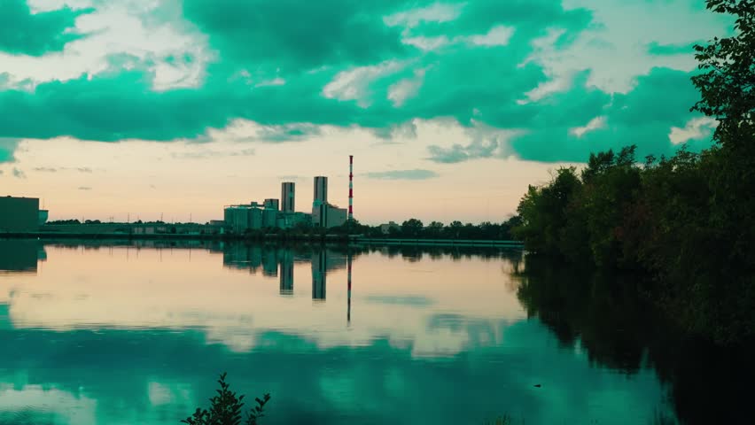 Industrial complex and smokestack mirrored on calm evening river, teal clouds and forested bank creating tranquil symmetry.