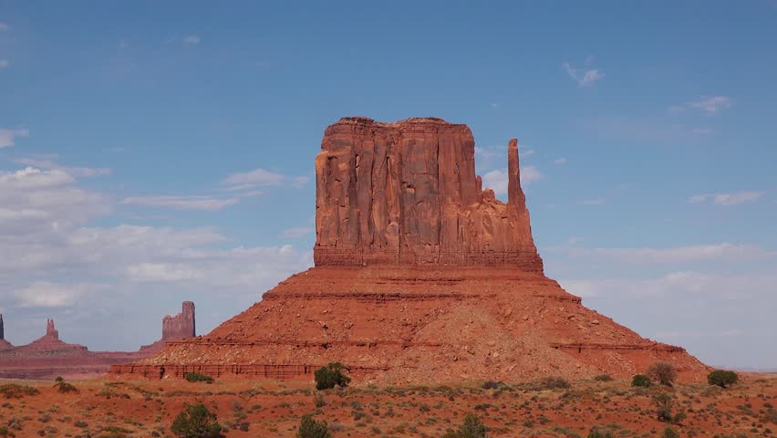 Majestic sandstone formations rise from the desert ground, surrounded by scattered vegetation. The sunlight illuminates the vibrant colors of the iconic landscape in Monument Valley.