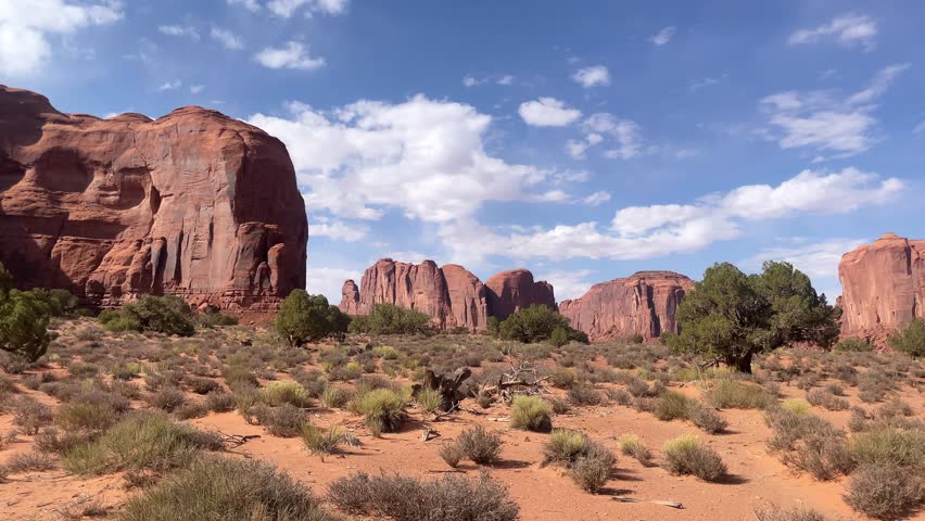 Majestic sandstone formations rise from the desert ground, surrounded by scattered vegetation. The sunlight illuminates the vibrant colors of the iconic landscape in Monument Valley.