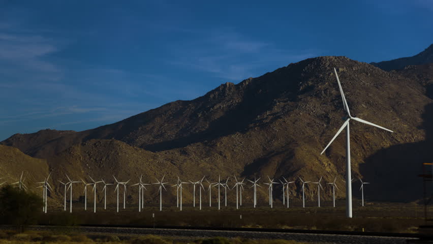Cinematic slow-motion passenger POV through the Southern California desert passing wind turbines turning in a field before a rugged mountain backdrop under a clear blue sky.