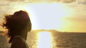 Pensive young woman with curly hair blowing in the wind watching a beautiful sunset over the ocean, with the bright sun reflecting on the water and creating a strong lens flare effect - Powered by Shutterstock - Get 15% off with code: PIKWIZARD15