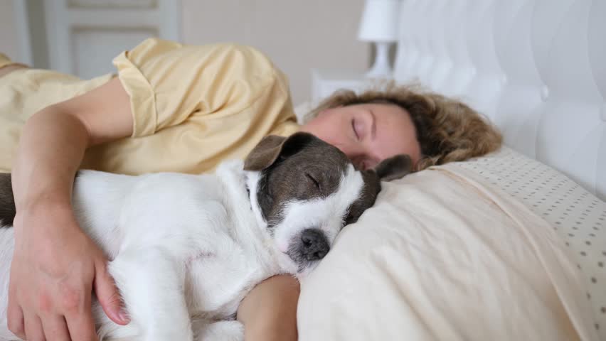 Peaceful moment of a young woman sleeping soundly in her bed while affectionately hugging her adorable puppy, sharing a comfortable and loving nap together in the serene light of the morning