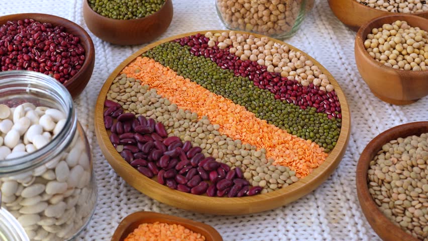 Various types of raw legumes, including beans, lentils, chickpeas, and soybeans, are beautifully arranged in wooden bowls and a central plate, highlighting a rich source of plant based protein