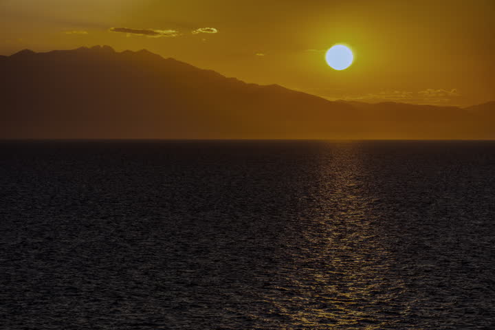 The sun is setting over the ocean, casting a warm glow on the water. The sky is filled with clouds, creating a serene and peaceful atmosphere in the Aegean sea in Greece