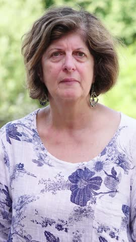 Vertical shot of an elderly woman with dark hair, looking seriously at the camera. Her intense gaze and serious expression are framed by the serene natural background, creating a powerful contrast.