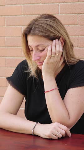 Vertical shot of a young woman with long blonde hair, sitting at a table and looking deeply sad. Her expression and posture suggest she is dealing with a recent breakup, 