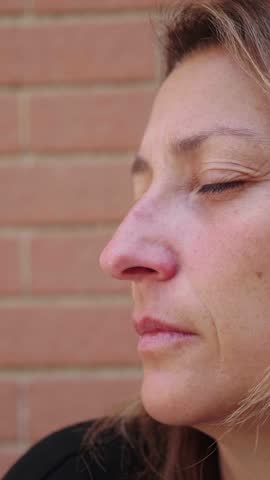 Vertical shot of a young woman with long blonde hair in profile, showing a contemplative and sad expression. Her eyes reflect deep thoughts and emotions, set against a blurred background.