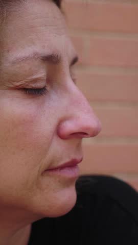 Vertical shot of a young woman with long blonde hair in profile, showing a contemplative and sad expression. Her eyes reflect deep thoughts and emotions, set against a blurred background.