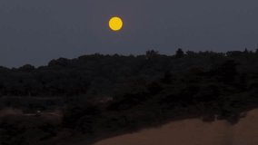 A large yellow full moon rises over a dark, silhouetted hill while a single white car drives along a winding road below. Telephoto shot capturing a moment of night travel - Powered by Shutterstock - Get 15% off with code: PIKWIZARD15
