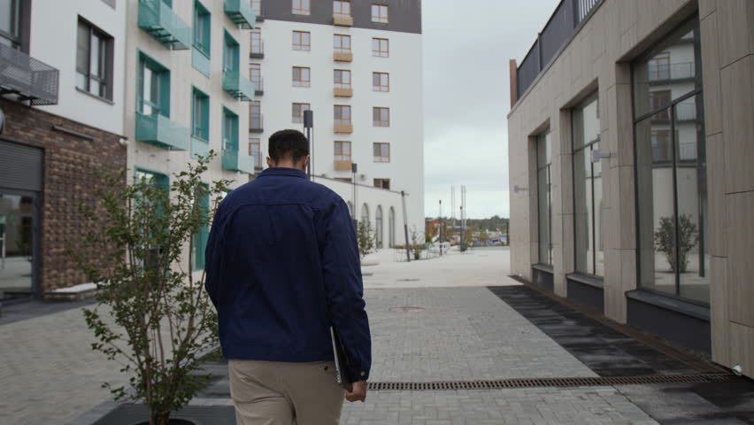 Back view of unrecognizable male programmer carrying laptop in hand while walking in street built up with modern buildings