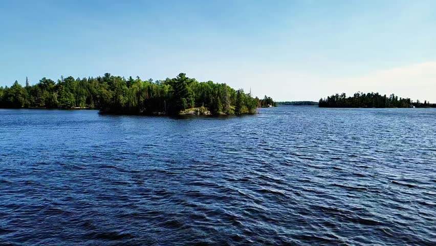 A peaceful scene of Kenora Lake in Ontario, Canada, captured on a calm, sunlit day. The deep blue water stretches toward the horizon, dotted with lush green islands beneath a clear summer sky. 