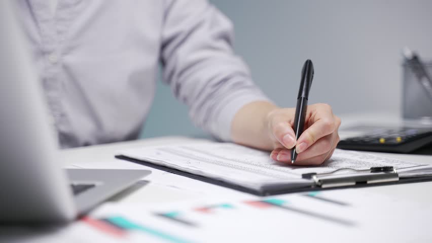 Close up of a female hand filling out documents with a pen at desk at workplace in a modern business office. Asian businesswoman is engaged in paperwork. Female accountant fills out a blank tax form