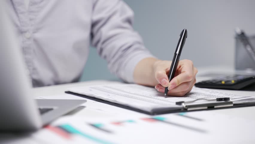 Close up of a female hand filling out documents with a pen at desk at workplace in a modern business office. Asian businesswoman is engaged in paperwork. Female accountant fills out a blank tax form