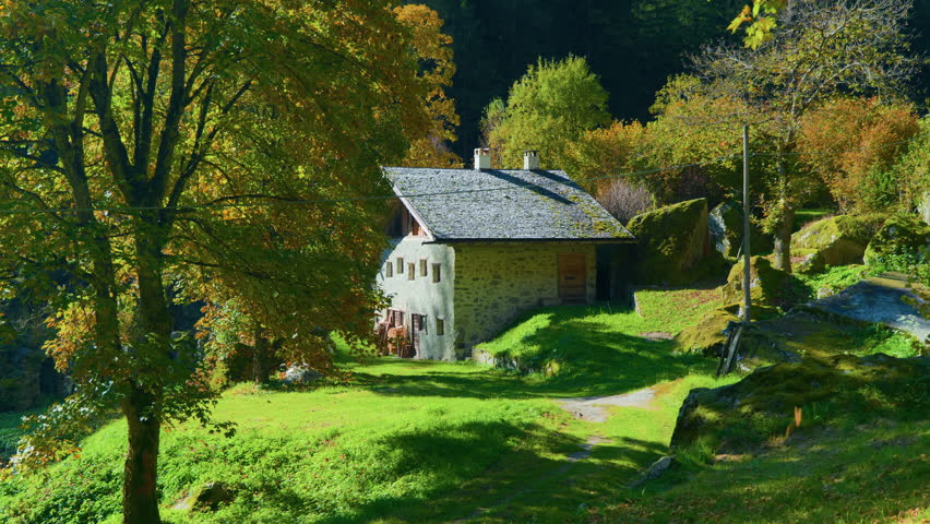 Wide shot of a small brick mountain cabin surrounded by a green meadow and forest, with touches of autumn-colored foliage.