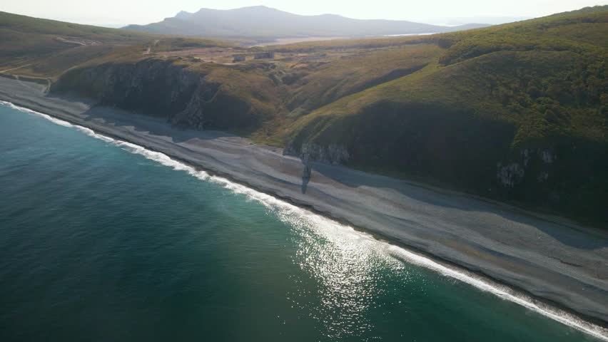 Aerial drone footage of the bay. A long pebble beach, turquoise waves, dramatic sea stacks and steep rocky cliffs along the wild Pacific coastline.
