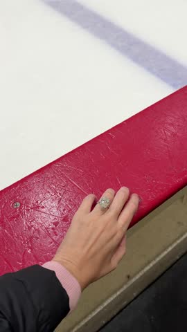 Woman’s hand with an oval diamond engagement ring placed on a red border at an indoor hockey rink with glossy white ice in the background.