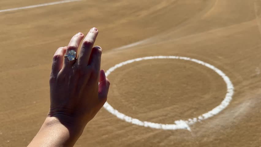 Closeup of a female hand with an oval diamond ring by a circle in the sandy surface of a baseball infield under clear bright sky.