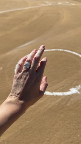 Woman’s hand wearing an oval diamond engagement ring placed on baseball field sand with white chalk lines and warm afternoon sunlight.