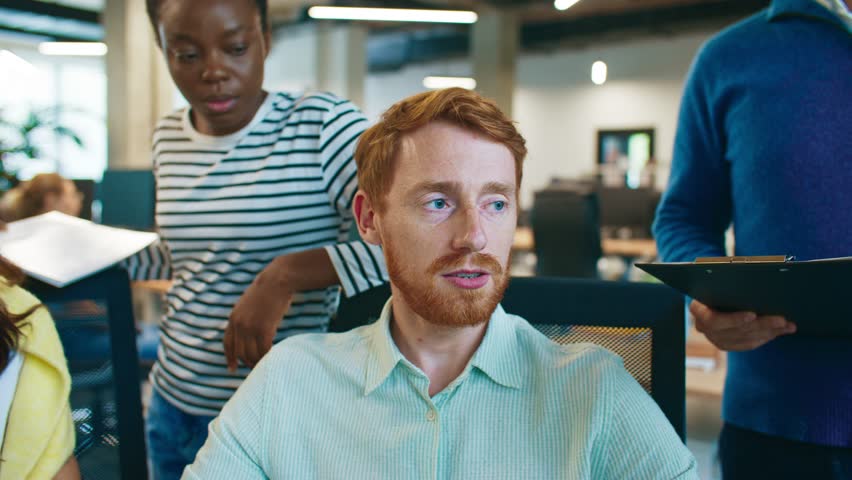 Team of diverse coworkers discussing project at workplace. Man pointing at computer screen explaining task while colleagues listening and taking notes. Group collaborating in modern office environment