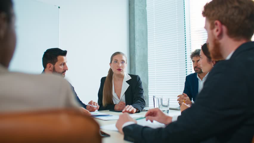 Elegant woman with glasses speaking during team meeting. Sitting at big table. Colleagues listening carefully and taking notes. Business discussion and strategy planning in modern conference room.
