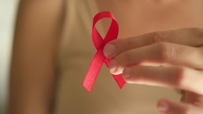 Close-up of female hand holding red satin ribbon, symbolizing HIV AIDS awareness, support for affected people, and hope for future cure.
