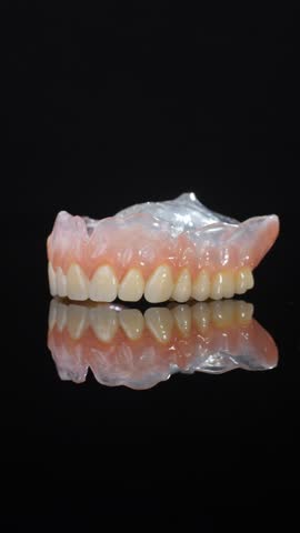 Removable denture shows natural teeth and pink gum base resting on black backdrop with reflection. Dental prosthesis reflects light while design shows precise modeling of jaw area and palatine part.