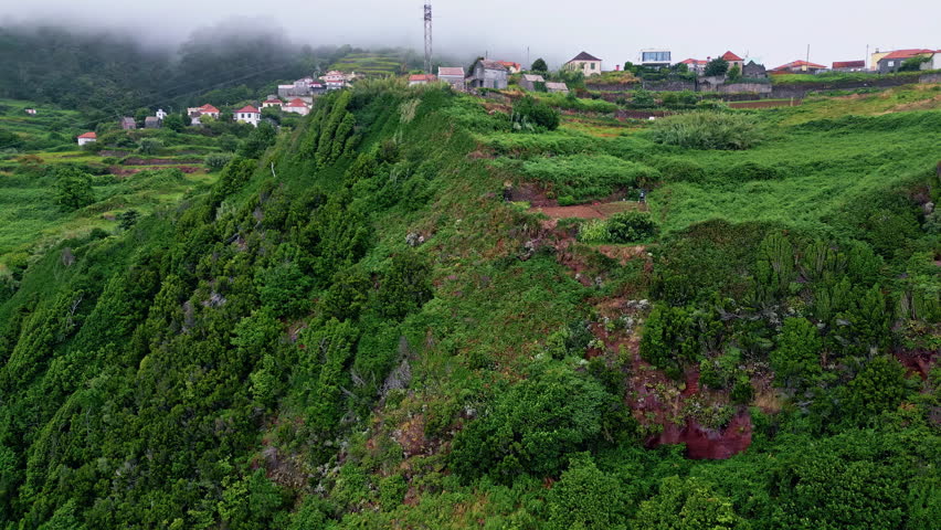 Village placed on green mountain slopes with lush fields and low clouds aerial view. Red-tiled houses create scenic rural landscape. Travel destination experiencing countryside living. Tourism concept