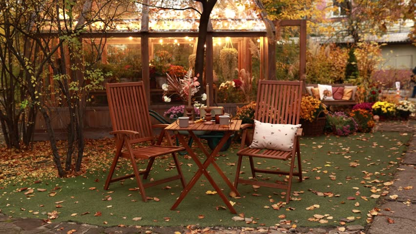 Exterior atmosphere in fall garden - table with flowers, cups, candles and wooden chairs with pillow for relax. Autumn yard with garden furniture set, decor and plants. Interior cozy patio at home.