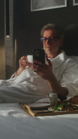 Man Enjoys Enjoys Breakfast in Bed and Phone in Hotel Room. Mature Man in Bathrobe Enjoys Leisurely Moment in Bed, Using Phone and Sipping Coffee. Concept Personal Time, Hotel Stay. Vertical Shot.