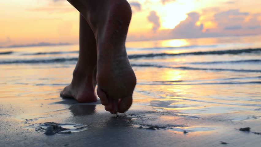 Silhouetted legs of a woman walking barefoot on a wet sandy beach during a beautiful sunset, with gentle sea waves washing over her feet and the warm evening light reflecting on the water