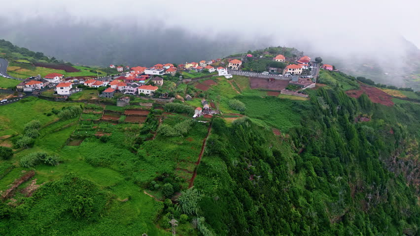 Lush island hills landscape drone shot. Breathtaking greenery mountain with low clouds sky on foggy morning. Tiny village houses on lush slopes. Rural area with scenic buildings. Picturesque nature