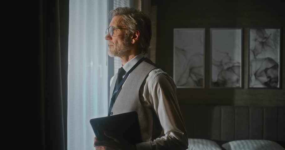 Executive Ready For Presentation In Sunny Hotel Room. Confident Businessman Stands In Sunlight By Window, Holding Digital Tablet, Ready For Meeting. Professional Training, Digital Workflow. Portrait.