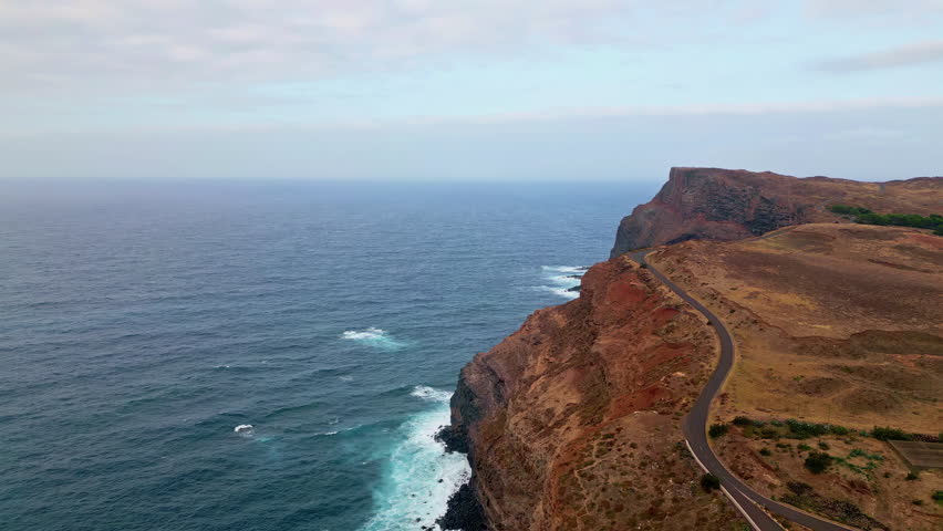 Stormy ocean crashing cliffs under cloudy sky. Picturesque panorama coastline with rough stone formations surrounded by sea water. Aerial view natural shoreline landscape. Road stretching seashore.