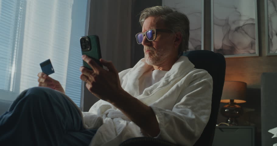 Gray Haired Man in Robe Sits by Window, Confirming Online Purchase With Phone and Credit Card. Concept of Modern Technology, Remote Work, Digital Finance, and Comfortable Hotel Living. Handheld.
