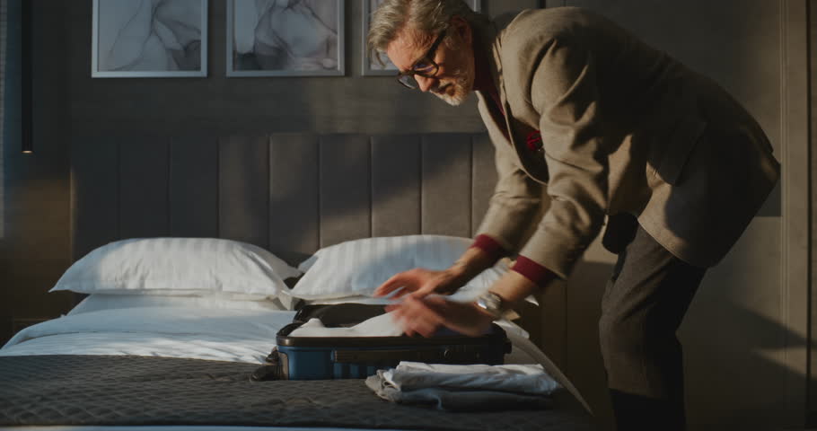 Elegant Gray Haired Man Meticulously Packs Clothes Into Small Blue Suitcase. Man Checking Out of Hotel in Hurry to Catch Flight. Concept of Travel, Routine of Traveler, Business Trip. Static Shot.
