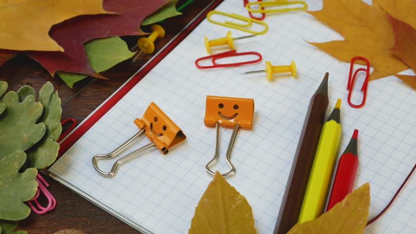 Autumn themed stationery arrangement with happy binder clips on notebook with cells, dried leaves. Vibrant office desk filled with stationery supplies. Back to school