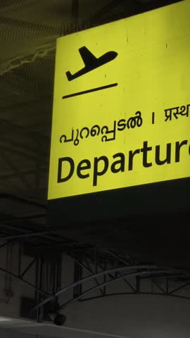 Vertical low-angle shot of yellow illuminated LED departure sign at international airport terminal, hanging from ceiling, guiding passengers toward boarding gates.