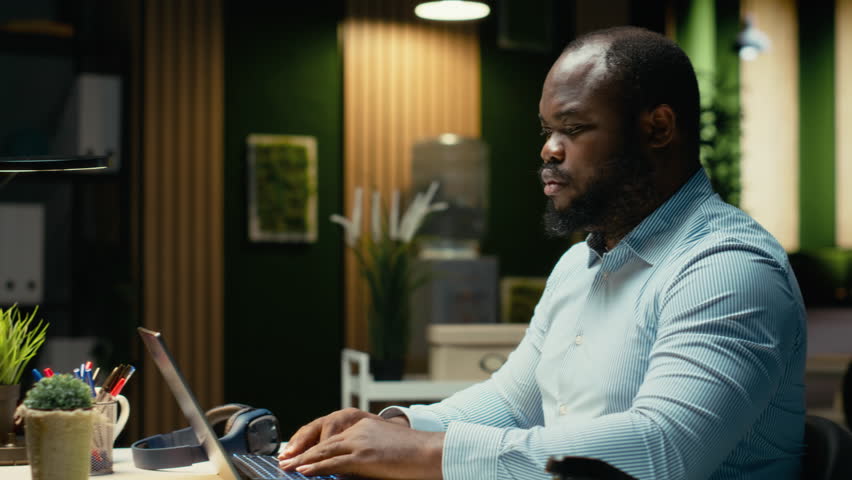 Black man consultant enjoying a cup of coffee to stay awake late at night, handling administrative tasks in the corporate space. Proofreading and writing at night with a caffeine drink. Camera B.