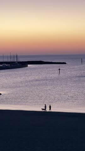 Romantic sunset scene at St Kilda Beach, Melbourne. People walking their dog along the calm shoreline under orange skies with distant boats and reflections on tranquil waters.