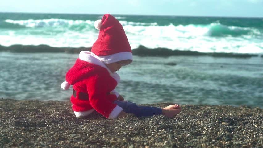 Child in Santa outfit playing on beach. Christmas vacation by the sea. Festive holiday mood in tropical setting. Cute child enjoying winter holiday warmth. Christmas joy near ocean waves.