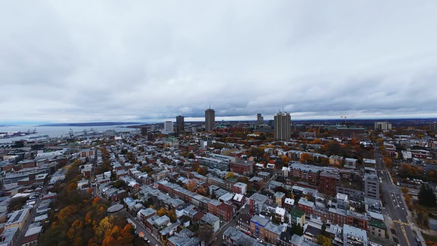 Low‑altitude 4K drone flyover toward Quebec City skyline—rows of houses, red roofs and bright fall trees along a hillside under clouds.