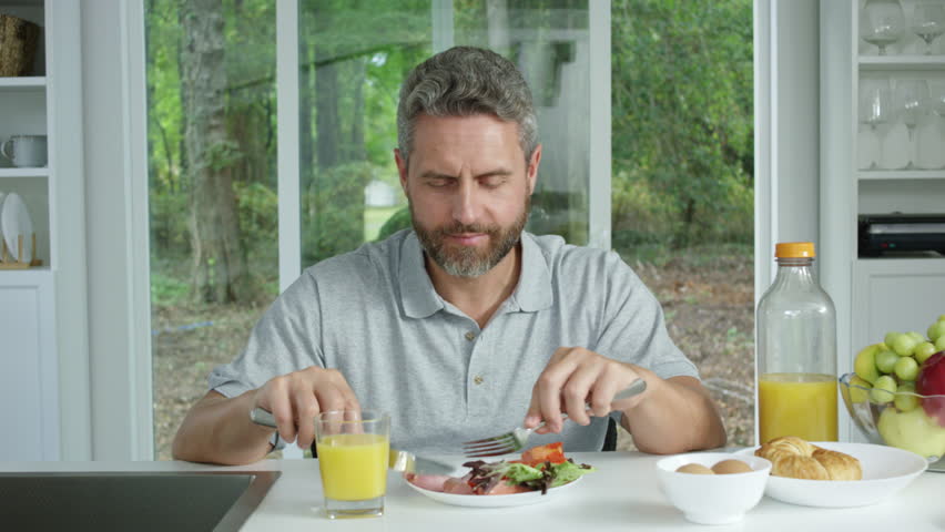 Man having healthy breakfast at home with fresh juice and salad. Mature adult eating nutritious meal in bright kitchen with natural light. Concept of balanced diet, wellness, and morning routine.