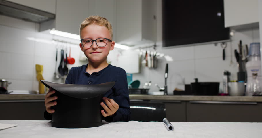 Happy boy wearing glasses playing magician in a modern kitchen, holding a black top hat with a white rabbit. A fun and imaginative moment creating magic and joy.