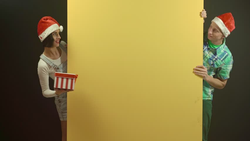 Playful Christmas scene with a man and woman wearing Santa hats behind a yellow wall. The woman offers a striped gift box with a smile. Couple fun on winter holiday concept.