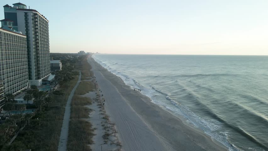 Aerial drone flying away from coastal buildings along Myrtle Beach, South Carolina, during sunrise golden hour.