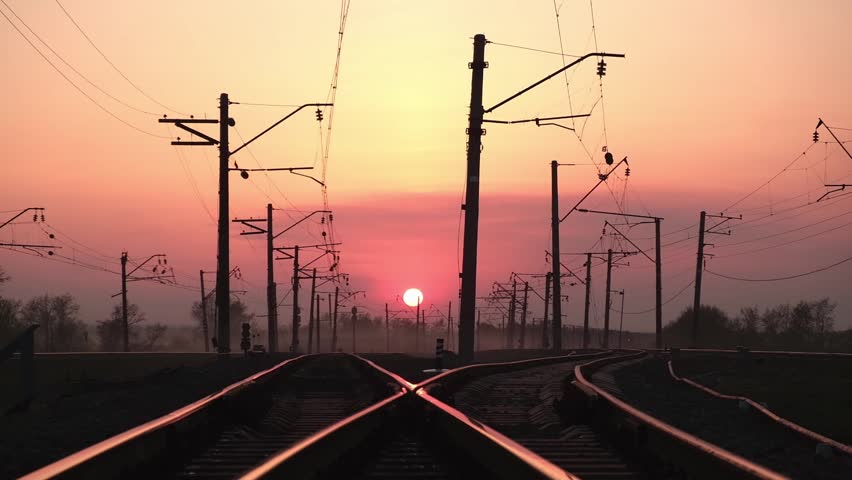 intersecting railway tracks leading towards horizon with vibrant pink and orange sunset in background, silhouetting electric poles and wires along route for scenic journey. close up.