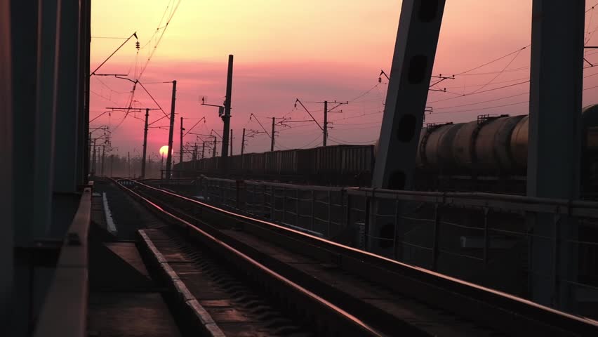 industrial landscape showing freight train with tank cars moving along railroad tracks over bridge during beautiful and colorful sunset, reflecting on steel rails. close up.
