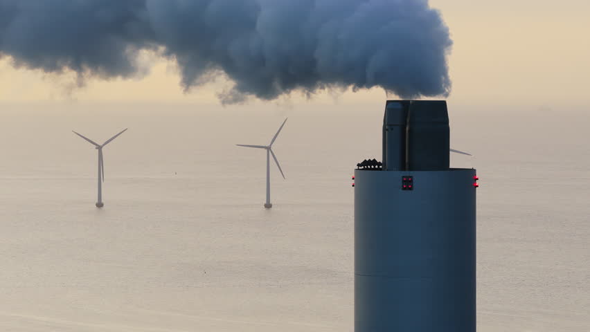 Aerial drone view of Amager Bakke power plant chimney releasing smoke, with offshore wind turbines standing in the sea behind it in Copenhagen, Denmark